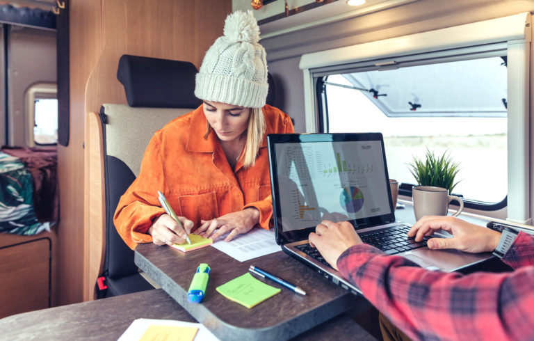 Woman in winter hat writing on sticky note inside RV with person on laptop across table